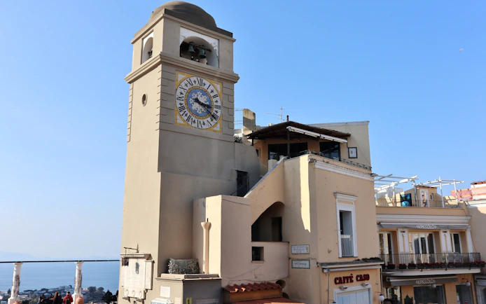 Clock tower in Piazza Umberto I, Capri, with nearby buildings and sea view.
