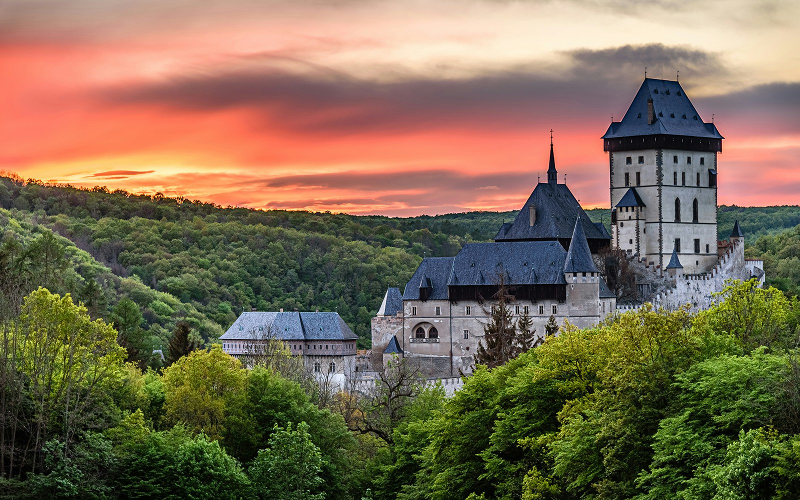 Karlštejn Castle surrounded by lush forest at sunset, showcasing Gothic architecture.