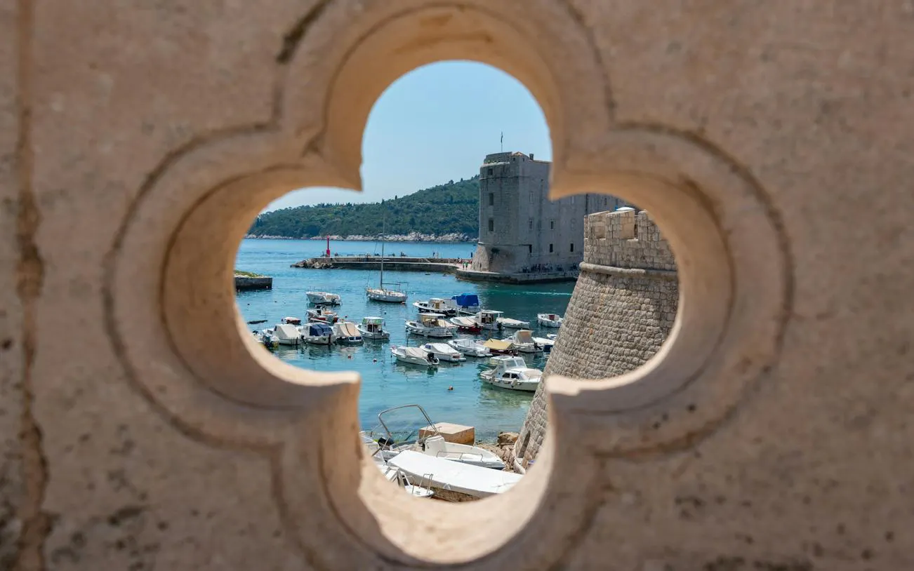 View of Dubrovnik Old Town harbor through stone wall opening, boats docked near fortress.