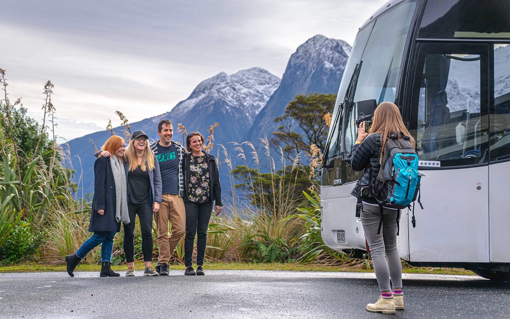 Group of passengers posing for a photo beside a coach with mountains in the background during a trip from Edinburgh to Loch Ness.