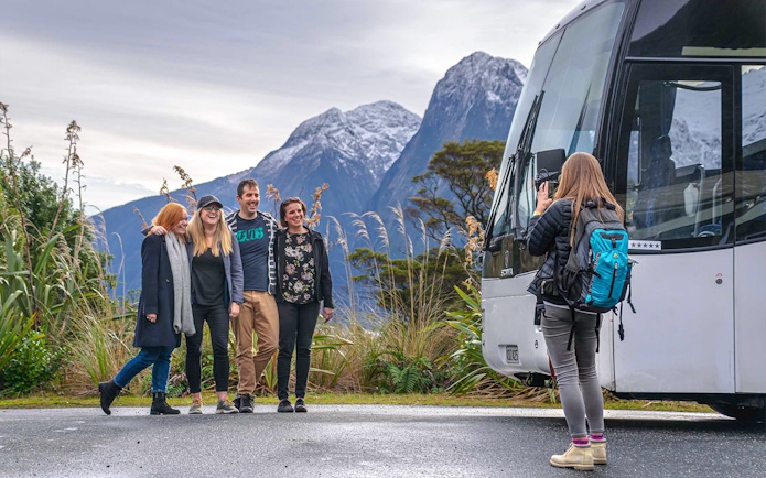 Group of passengers posing for a photo beside a coach with mountains in the background during a trip from Edinburgh to Loch Ness.