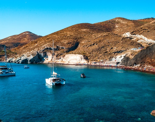 Catamarans anchored near Santorini's Red Beach with rocky hills in the background.