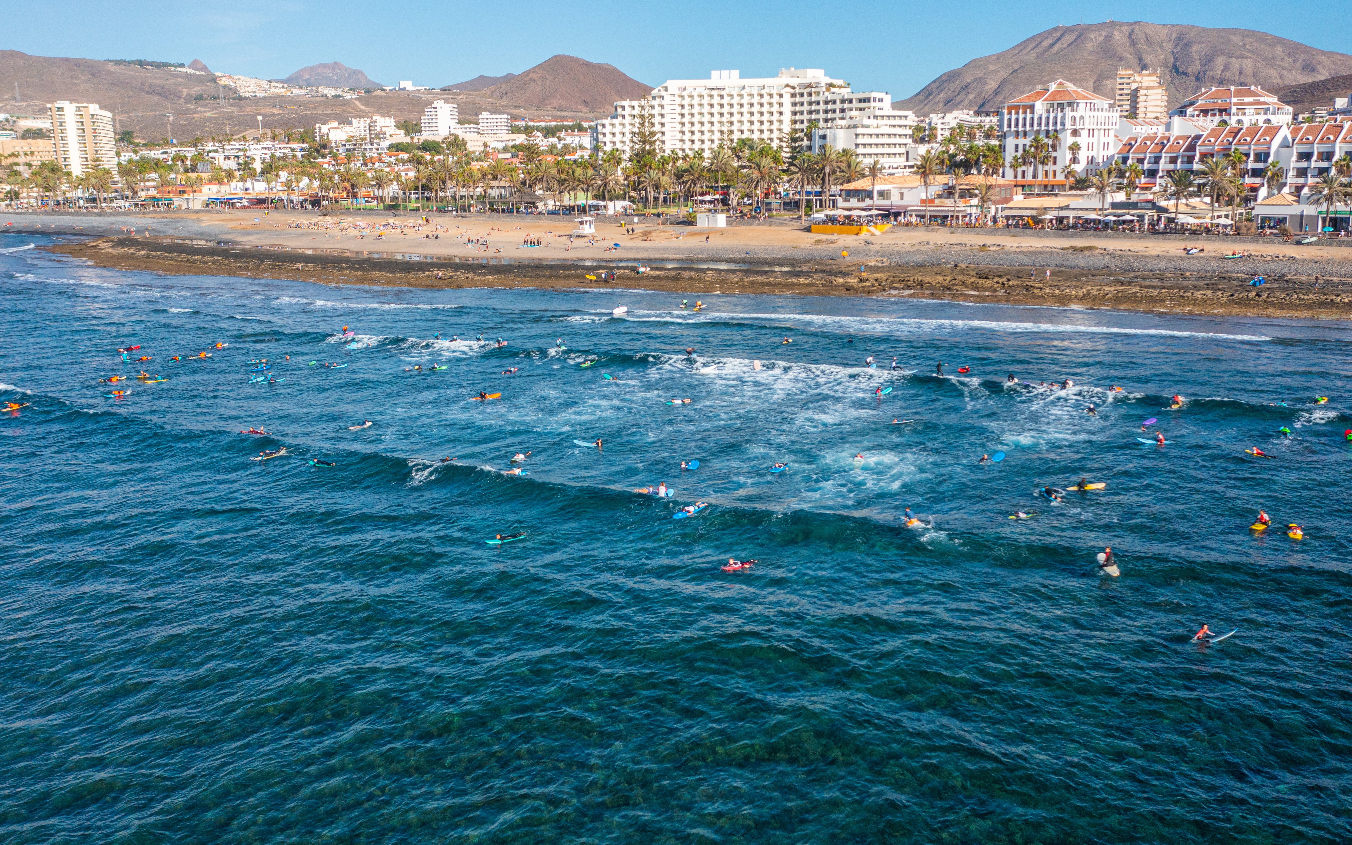 Surfers riding waves at Playa de las Americas, Adeje, with beachfront and mountains in the background.