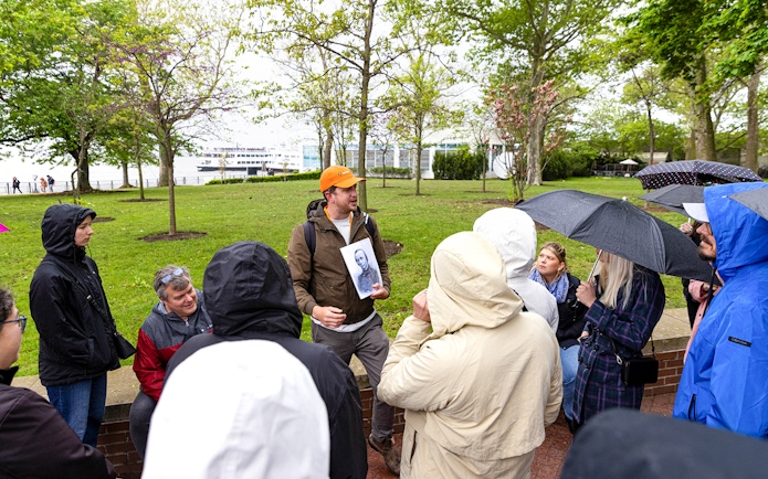 Guide speaking to visitors by the Wall of Honor on Ellis Island, holding a historical photo.