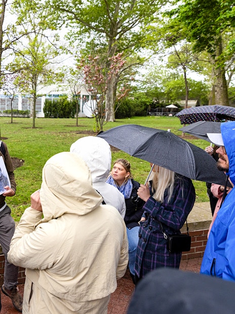 Guide speaking to visitors by the Wall of Honor on Ellis Island, holding a historical photo.