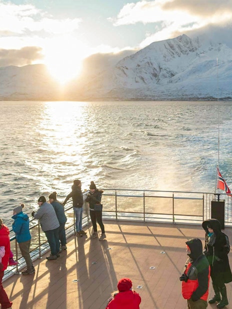 Tourists on a hybrid-electric boat in Tromso, Norway, with snowy mountains and sunset in the background.