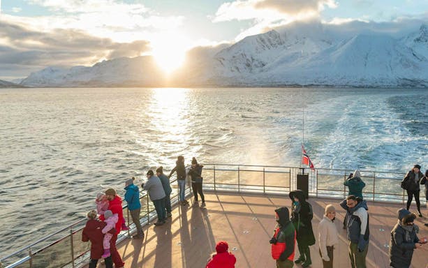 Tourists on a hybrid-electric boat in Tromso, Norway, with snowy mountains and sunset in the background.