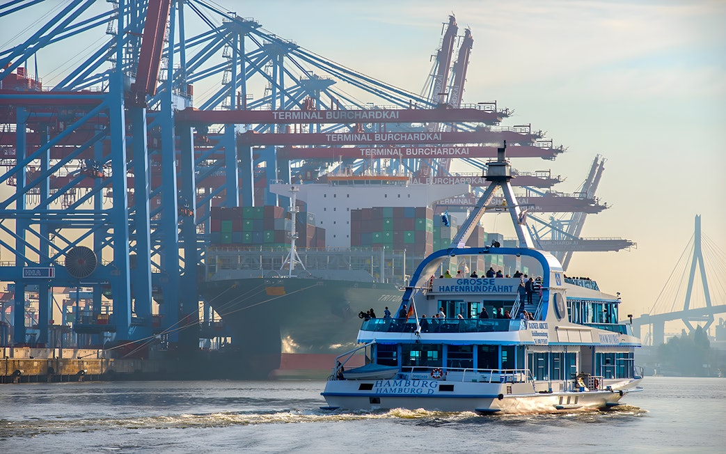 Hamburg harbor cruise ship passing container terminal cranes.