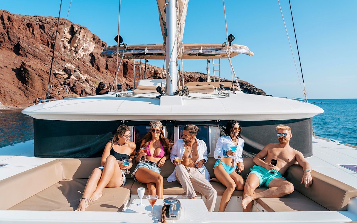 Group enjoying drinks on a catamaran cruise near Santorini's rocky coastline.