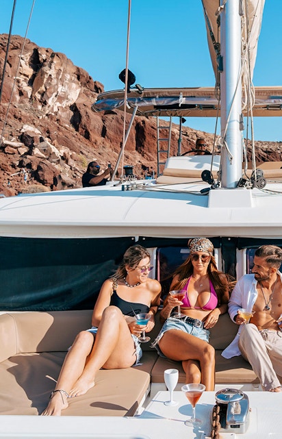 Group enjoying drinks on a catamaran cruise near Santorini's rocky coastline.