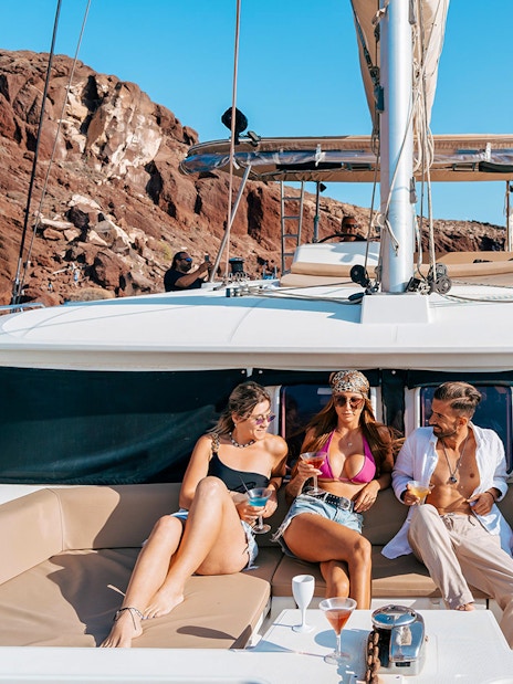 Group enjoying drinks on a catamaran cruise near Santorini's rocky coastline.