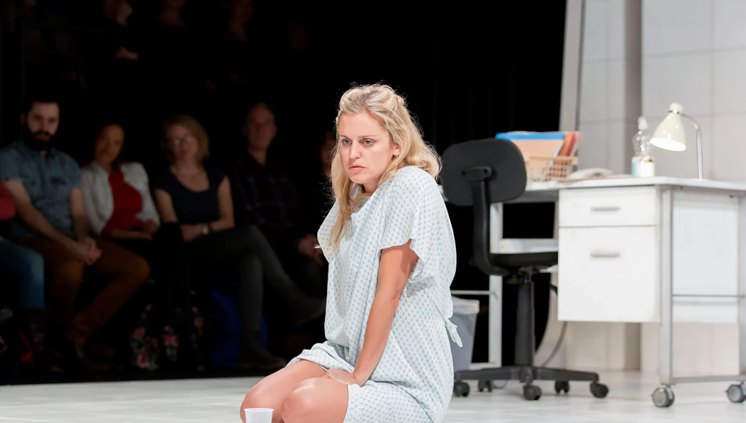 A woman in a hospital gown sits on the floor of a clinical setting with an audience watching.