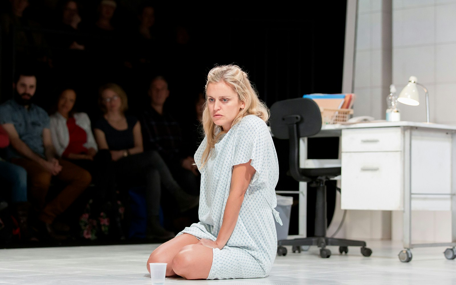 A woman in a hospital gown sits on the floor of a clinical setting with an audience watching.