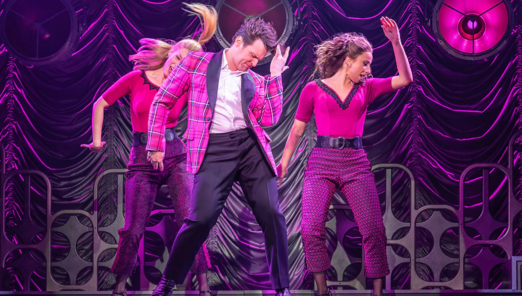 Performers dancing energetically on stage in a vibrant pink setting.