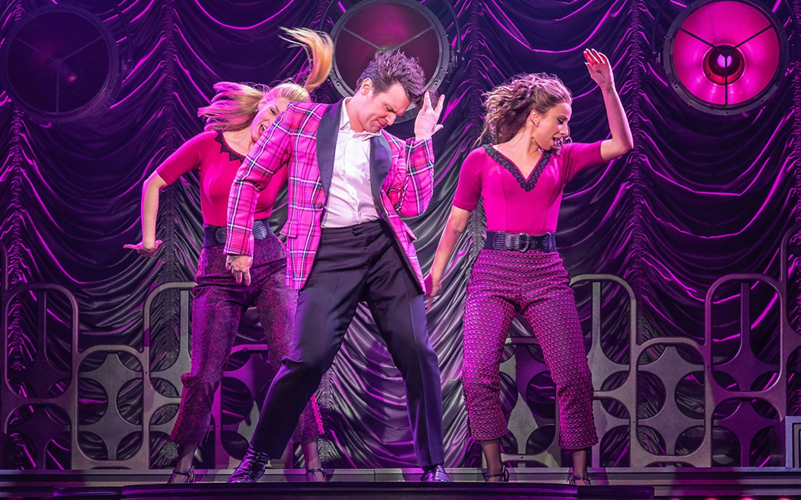 Performers dancing energetically on stage in a vibrant pink setting.