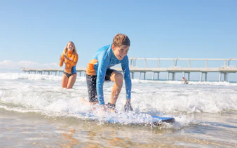 Beginner surf lesson for teens near a pier with instructor guiding in the background.