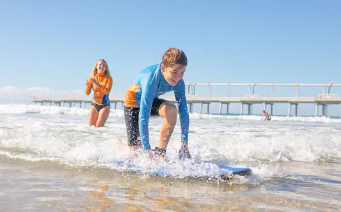 Beginner surf lesson for teens near a pier with instructor guiding in the background.