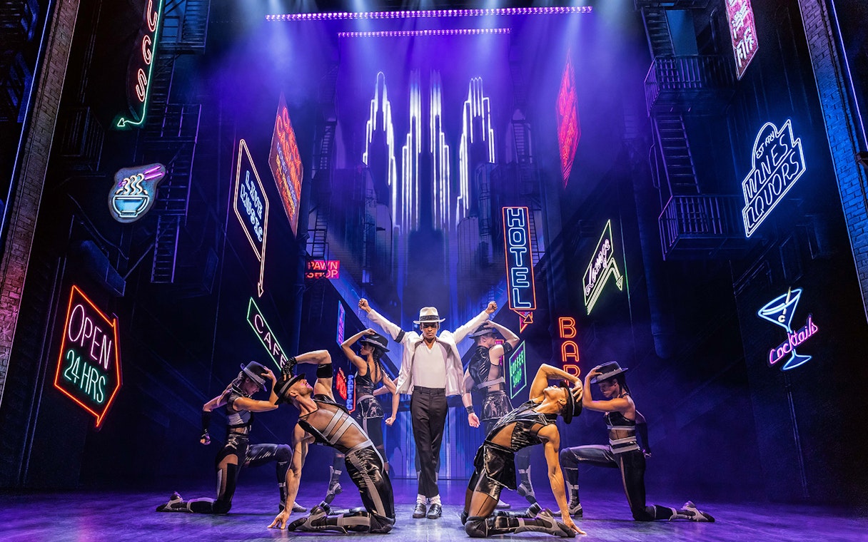 Performer in white hat and dancers on stage with neon city backdrop in MJ The Musical.