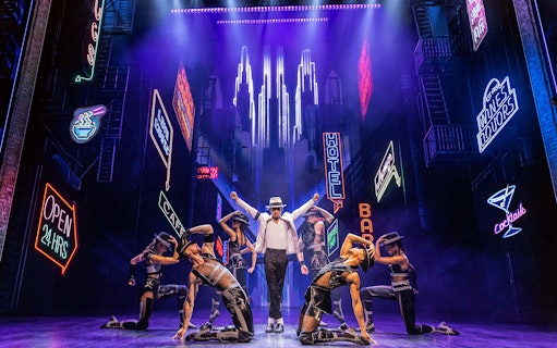 Performer in white hat and dancers on stage with neon city backdrop in MJ The Musical.