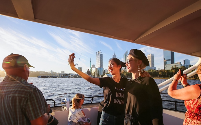 Tourists taking a selfie on a boat during sunset with city skyline in the background.