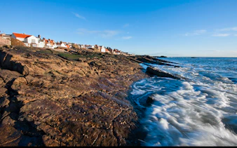 Coastal view of fishing village in Fife, Scotland, with rocky shoreline and ocean waves.