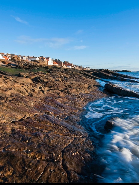 Coastal view of fishing village in Fife, Scotland, with rocky shoreline and ocean waves.