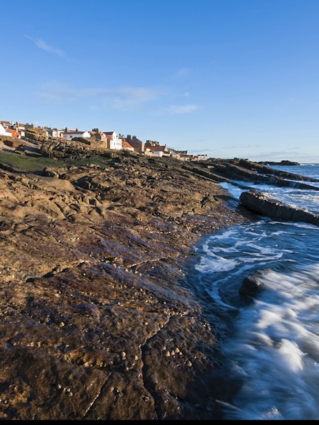Coastal view of fishing village in Fife, Scotland, with rocky shoreline and ocean waves.