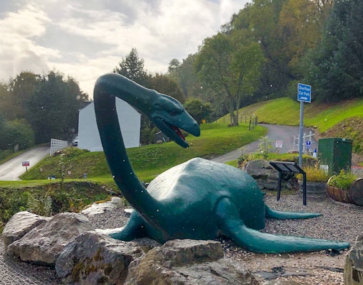 Loch Ness monster statue near a road and greenery in Scotland.