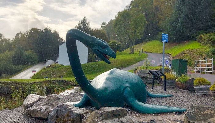 Loch Ness monster statue near a road and greenery in Scotland.