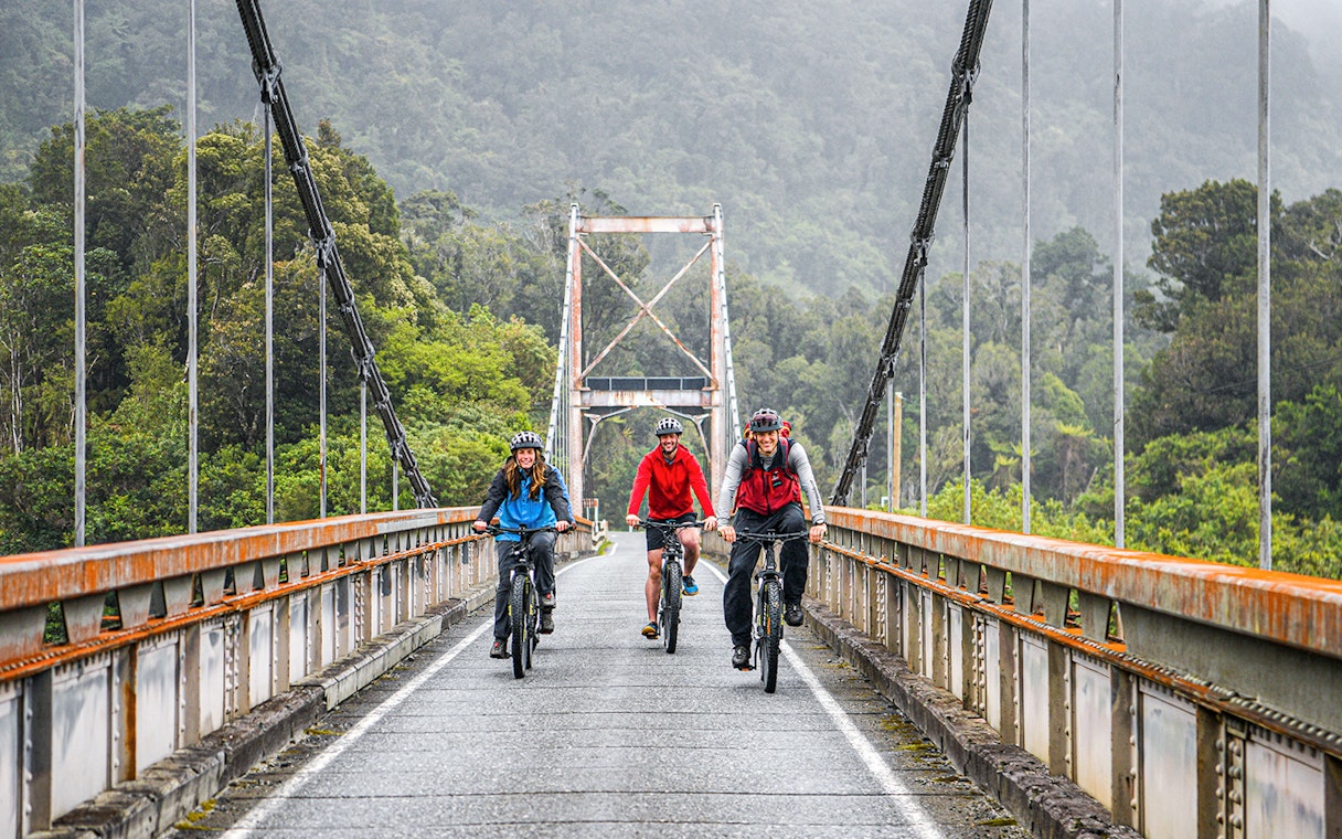 Tour group biking on a bridge in Fox Glacier Valley, surrounded by lush greenery.