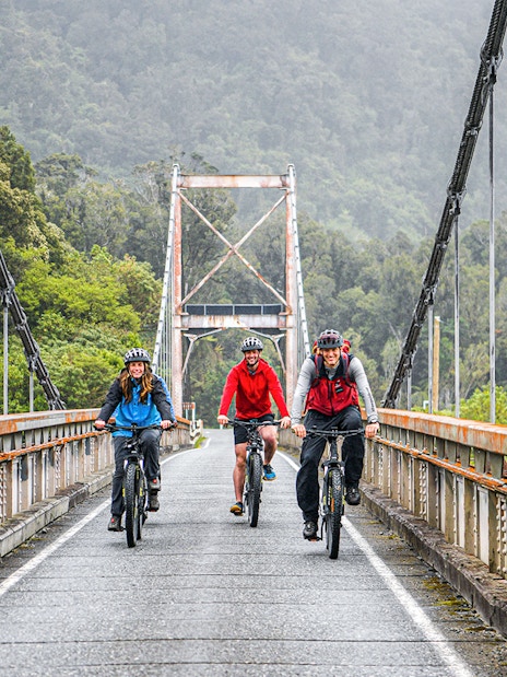 Tour group biking on a bridge in Fox Glacier Valley, surrounded by lush greenery.