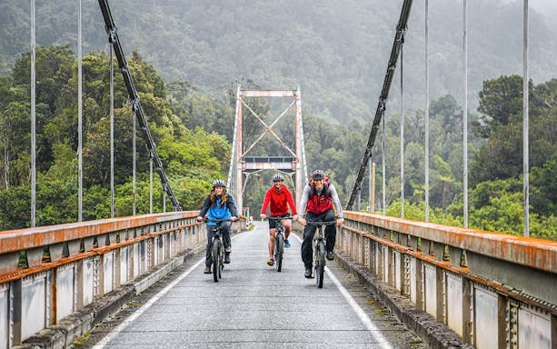 Tour group biking on a bridge in Fox Glacier Valley, surrounded by lush greenery.