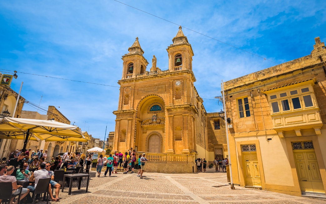 Tourists at a café near a historic church in Marsaxlokk village, Malta.