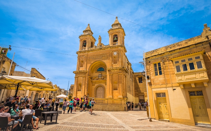 Tourists at a café near a historic church in Marsaxlokk village, Malta.