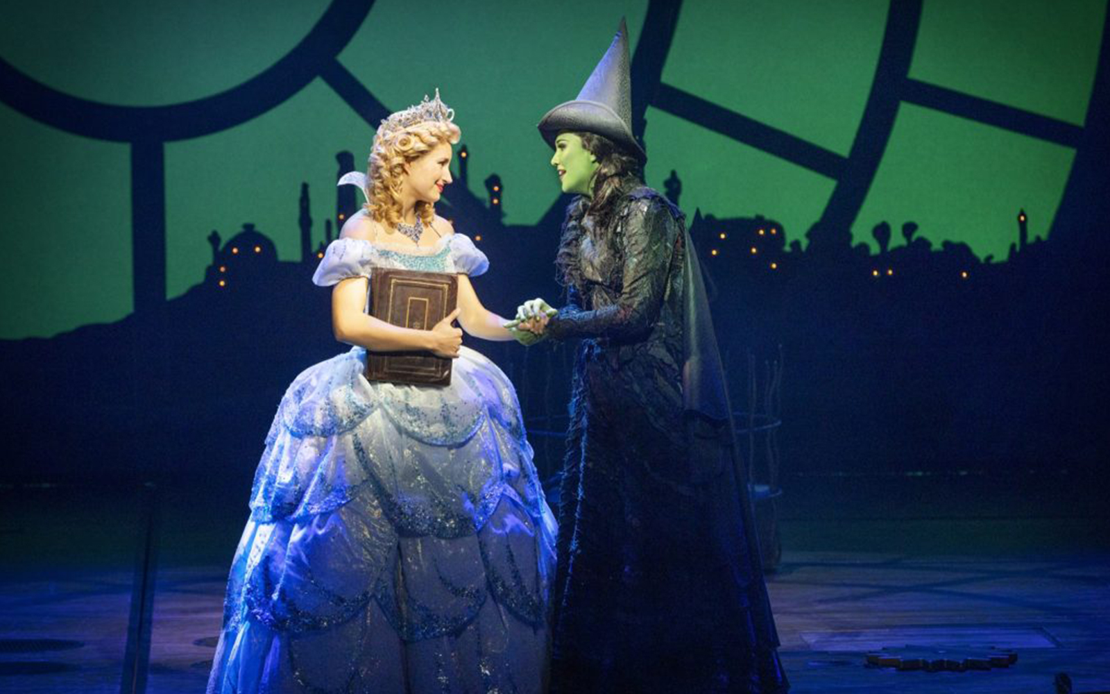 Two characters from Wicked the Musical on stage, one in a blue gown holding a book, the other in a black hat.
