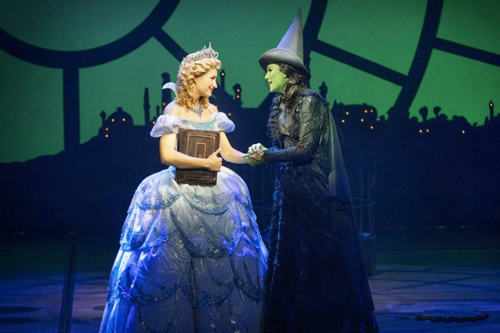 Two characters from Wicked the Musical on stage, one in a blue gown holding a book, the other in a black hat.