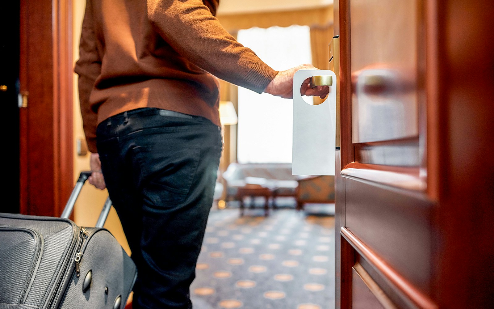 Person entering hotel room with suitcase, door hanger visible.
