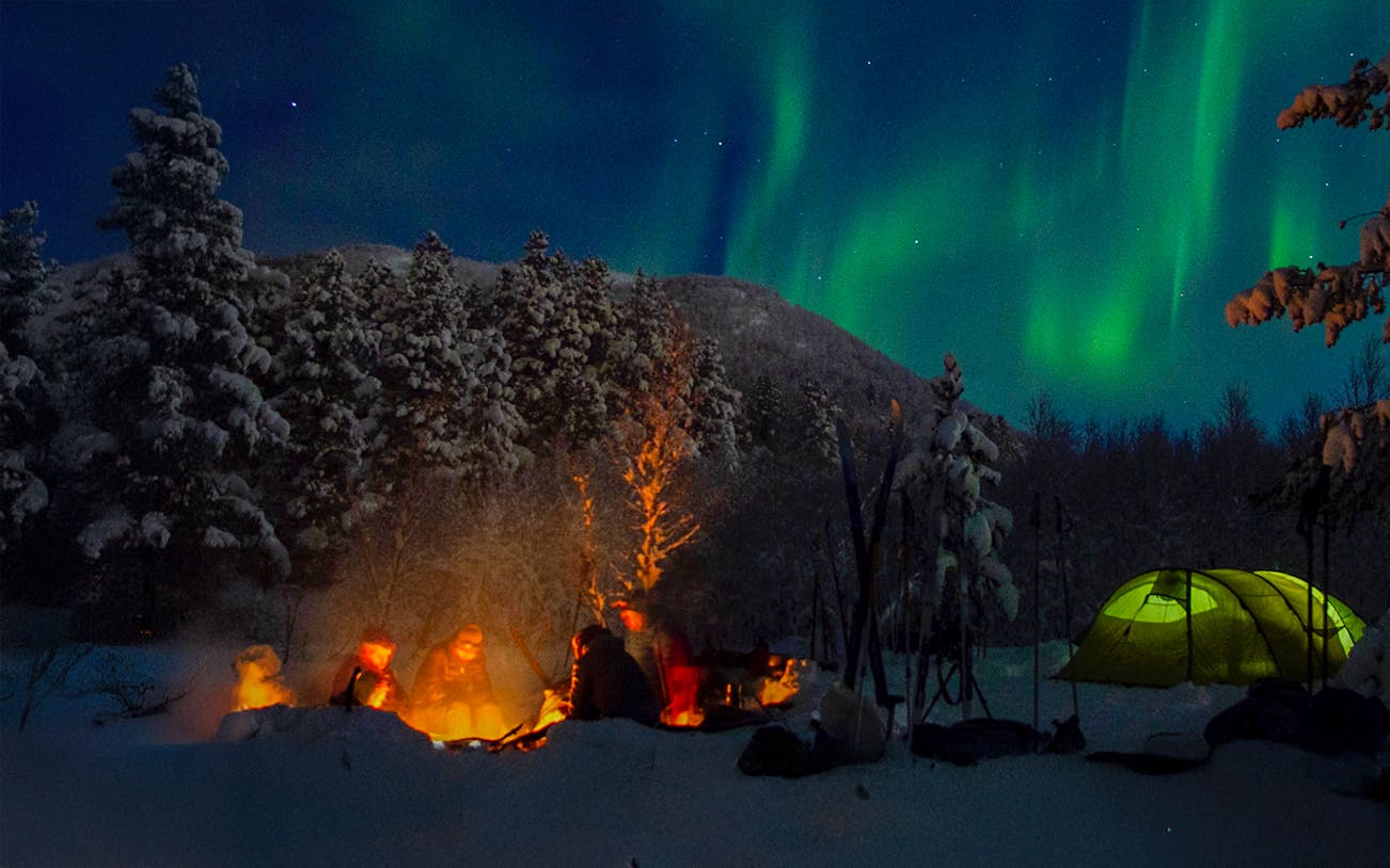 Campfire gathering under Northern Lights during a Lappish barbecue in snowy forest.