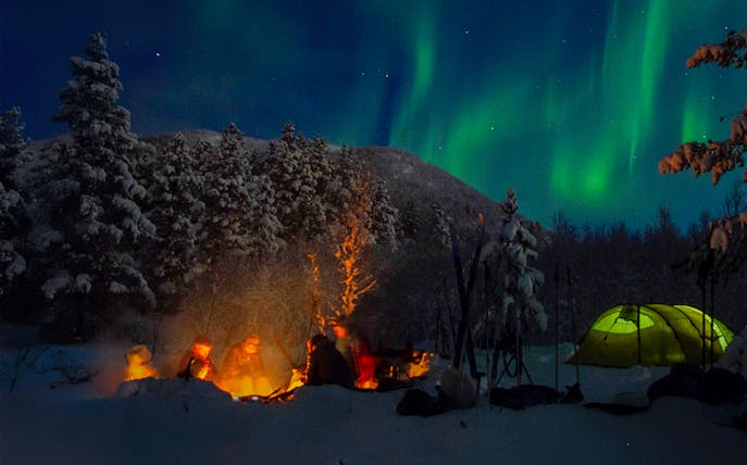 Campfire gathering under Northern Lights during a Lappish barbecue in snowy forest.