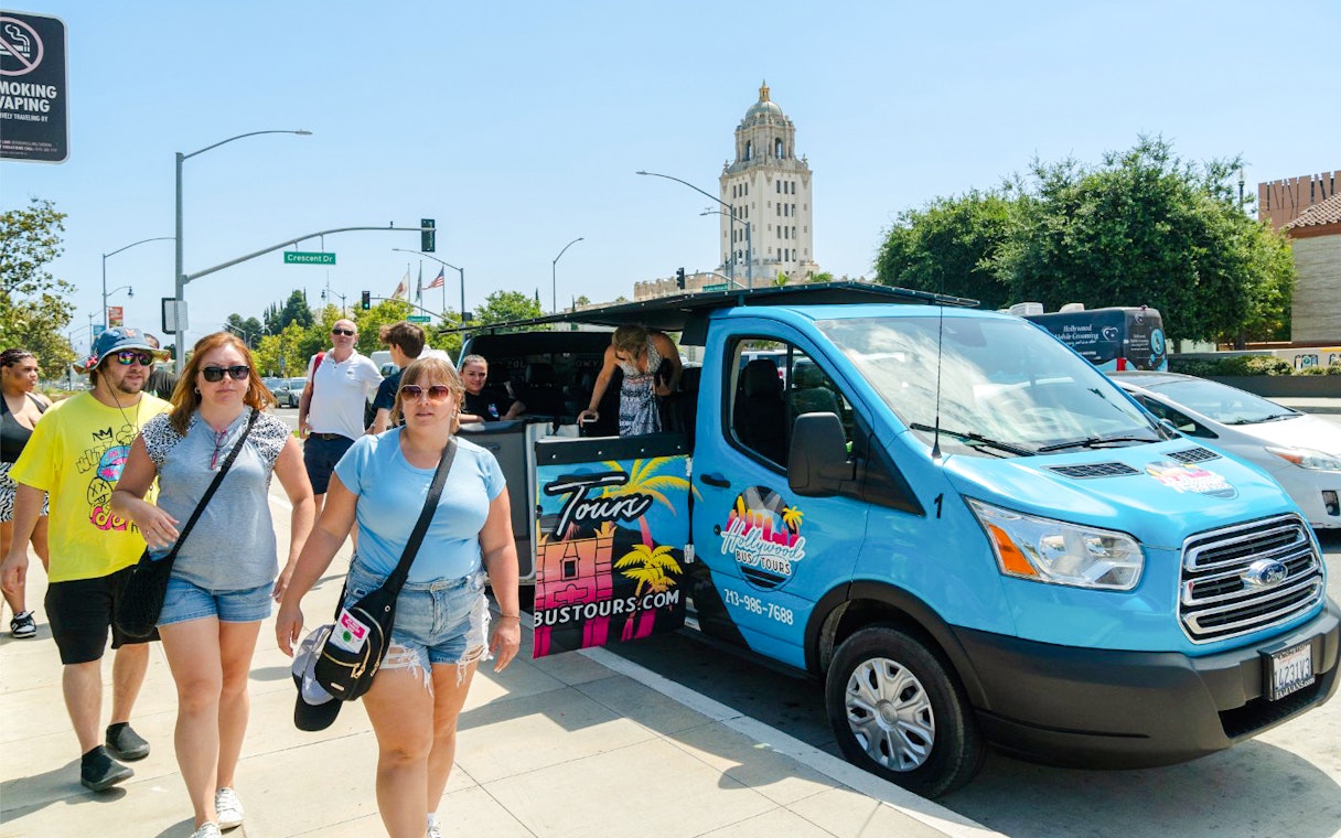 Tourists exiting a Celebrity Homes tour bus in Los Angeles with Beverly Hills building in background.