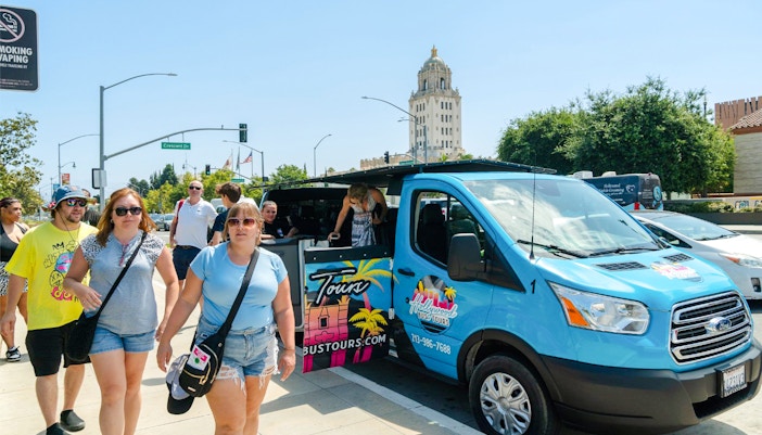 Tourists exiting a Celebrity Homes tour bus in Los Angeles with Beverly Hills building in background.