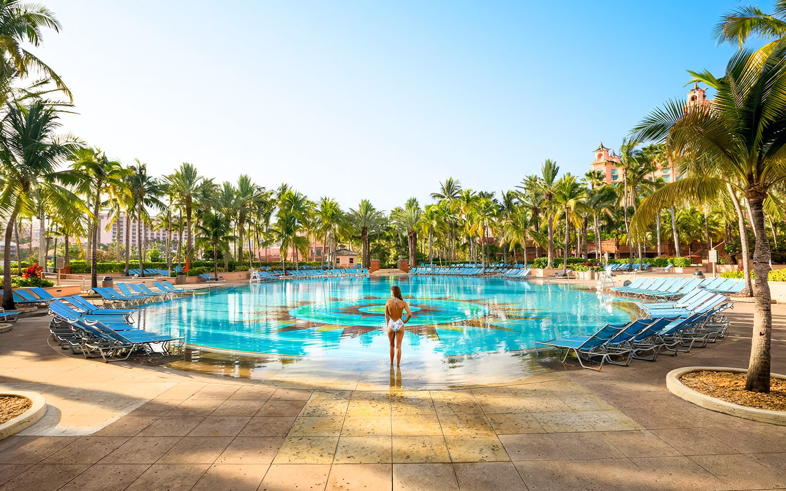 Person standing at the edge of a large pool surrounded by palm trees at Atlantis Aquaventure, Nassau, Bahamas.