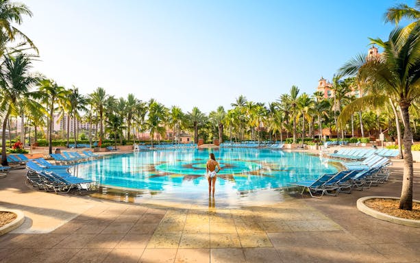 Person standing at the edge of a large pool surrounded by palm trees at Atlantis Aquaventure, Nassau, Bahamas.
