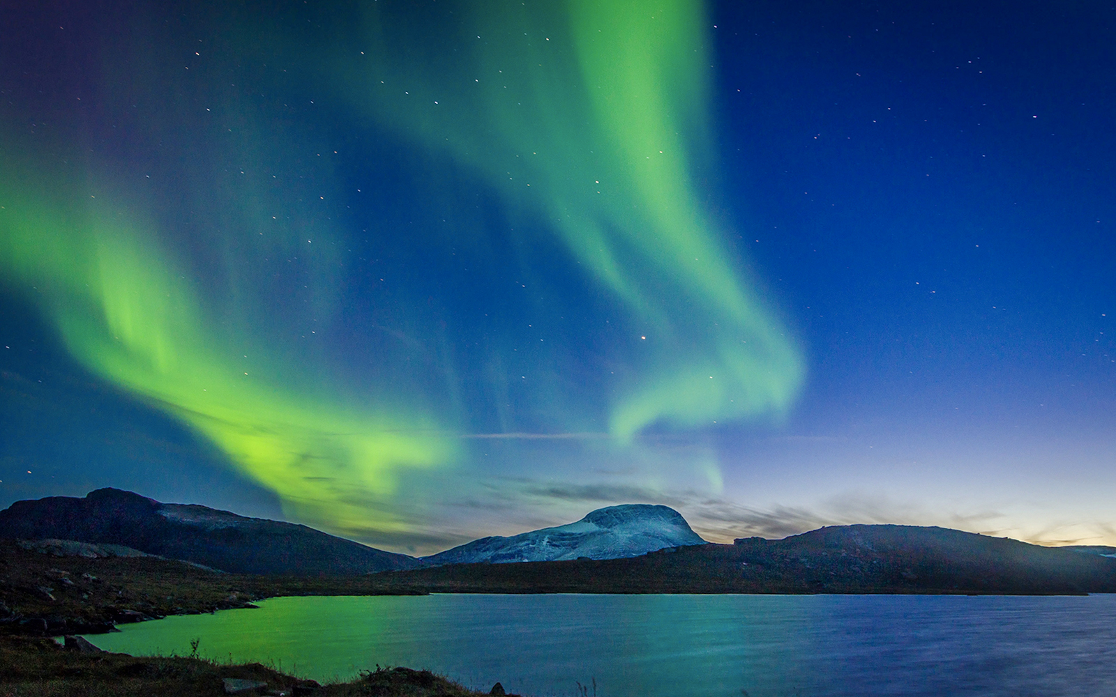 Northern lights over a mountain landscape with a lake in the foreground.
