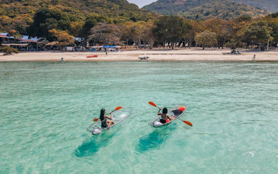 Kayakers in clear canoes on turquoise water near Pattaya beach with forested hills in the background.