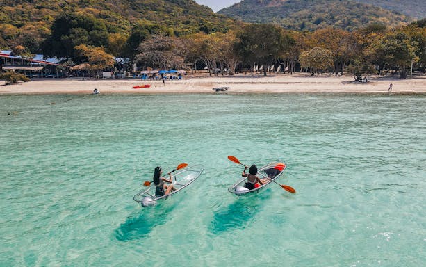 Kayakers in clear canoes on turquoise water near Pattaya beach with forested hills in the background.