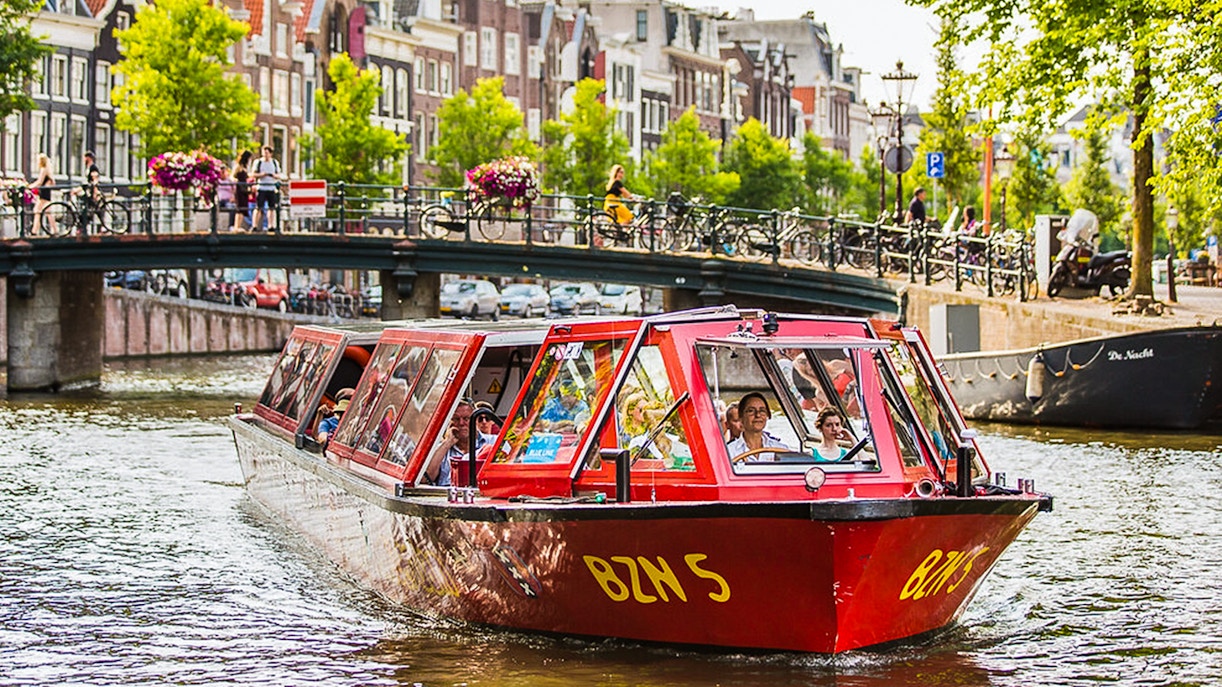 Hop-on hop-off boat on Amsterdam canal with bridge and historic buildings in background.