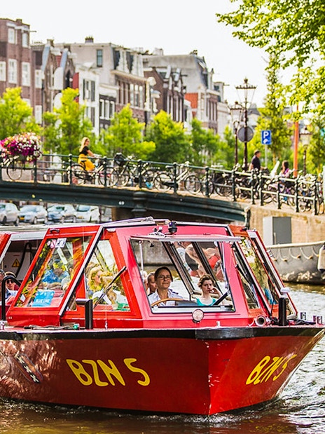 Hop-on hop-off boat on Amsterdam canal with bridge and historic buildings in background.