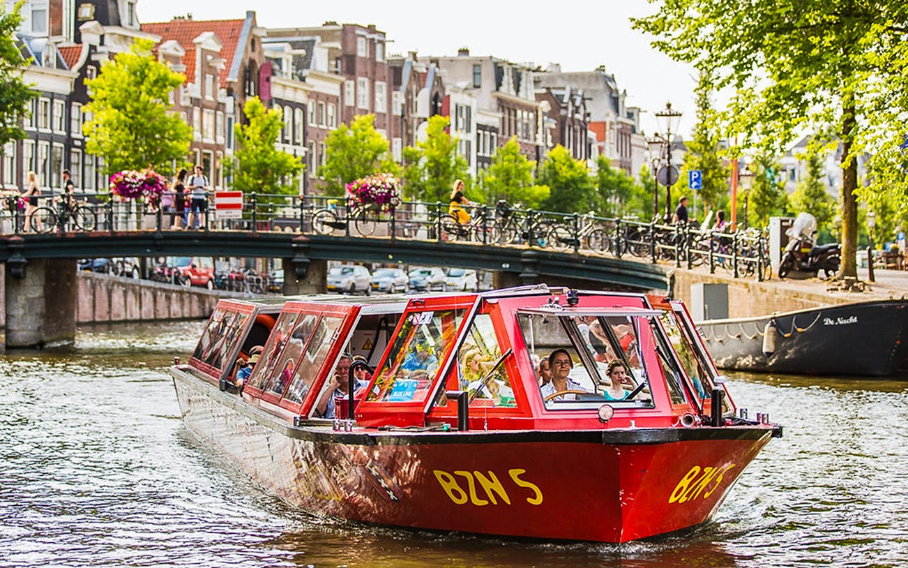 Hop-on hop-off boat on Amsterdam canal with bridge and historic buildings in background.