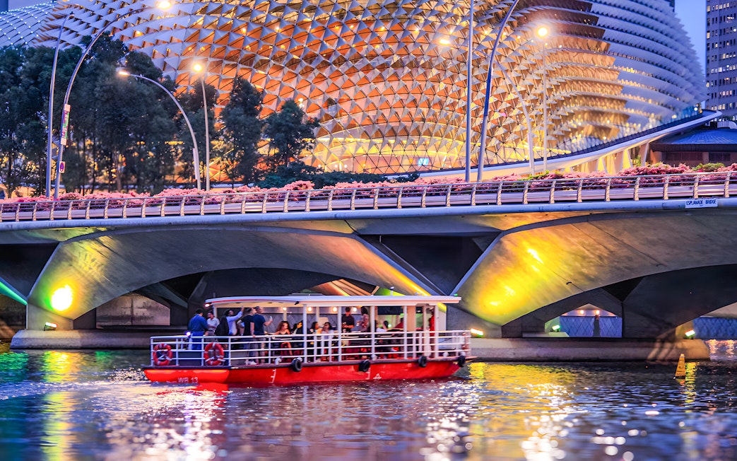 Singapore River Cruise boat passing under Esplanade Bridge at dusk.